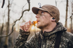 A hunter wears the First Lite-Stormy Kromer cap while using an animal hunting call in the woods.