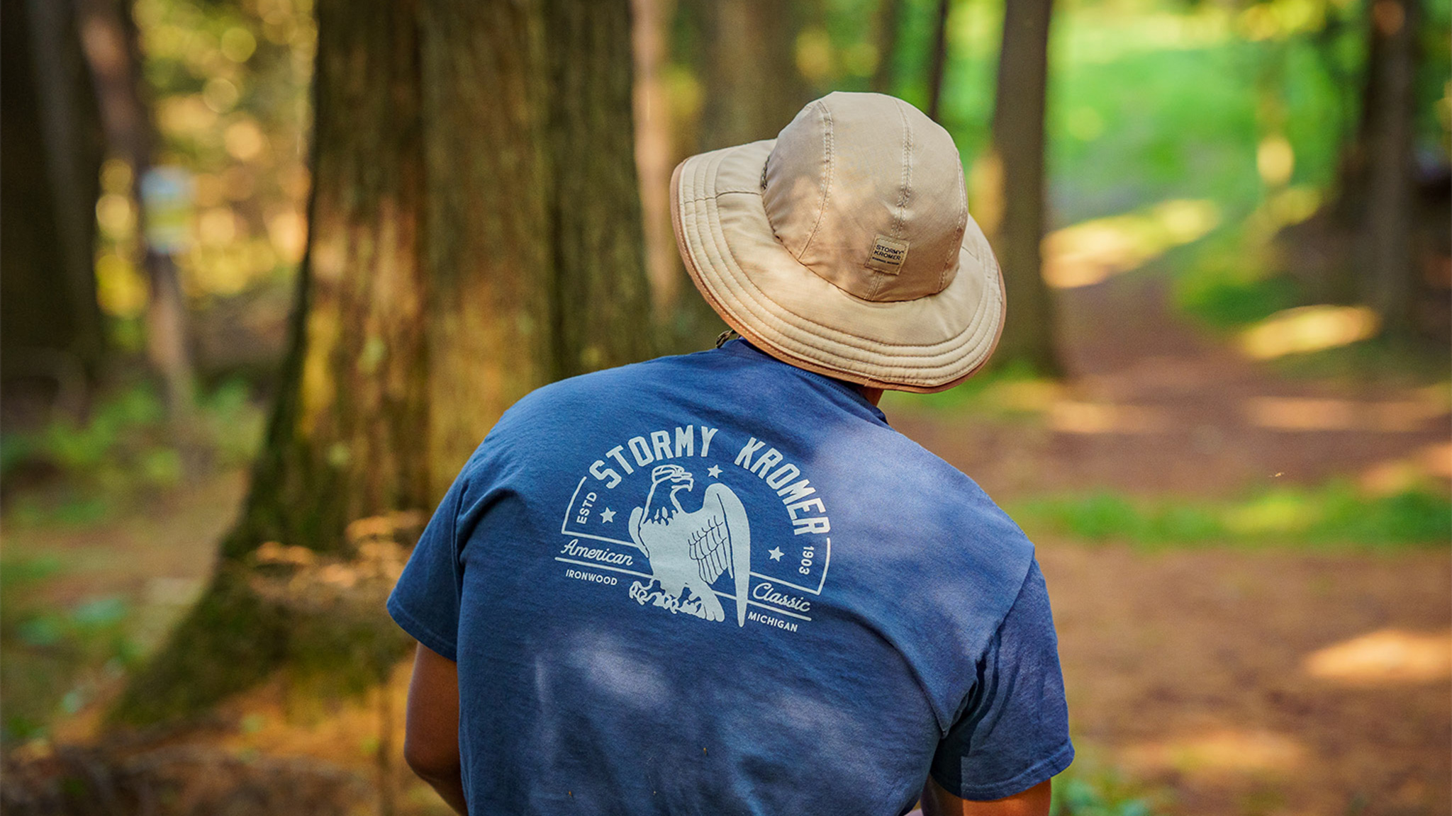 SK The Forager Hat Spring Launch a person sitting down in the wood with their back to the camera wearing a stormy kromer hat and tshirt