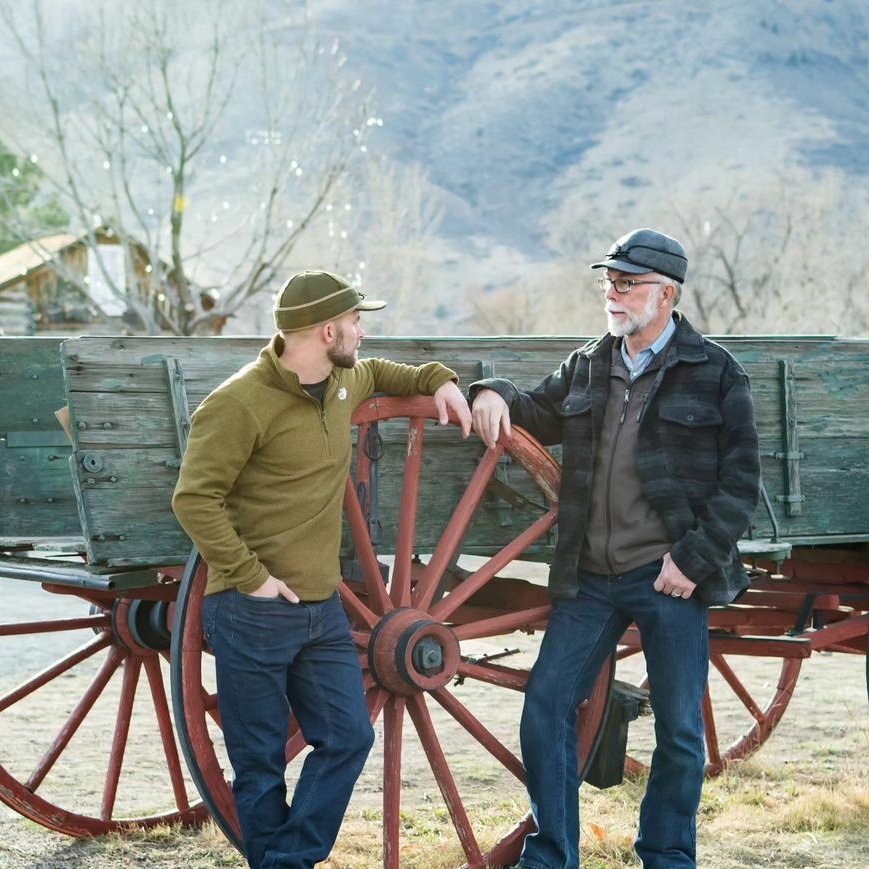 Two men lean against a wagon while wearing Stormy Kromer attire