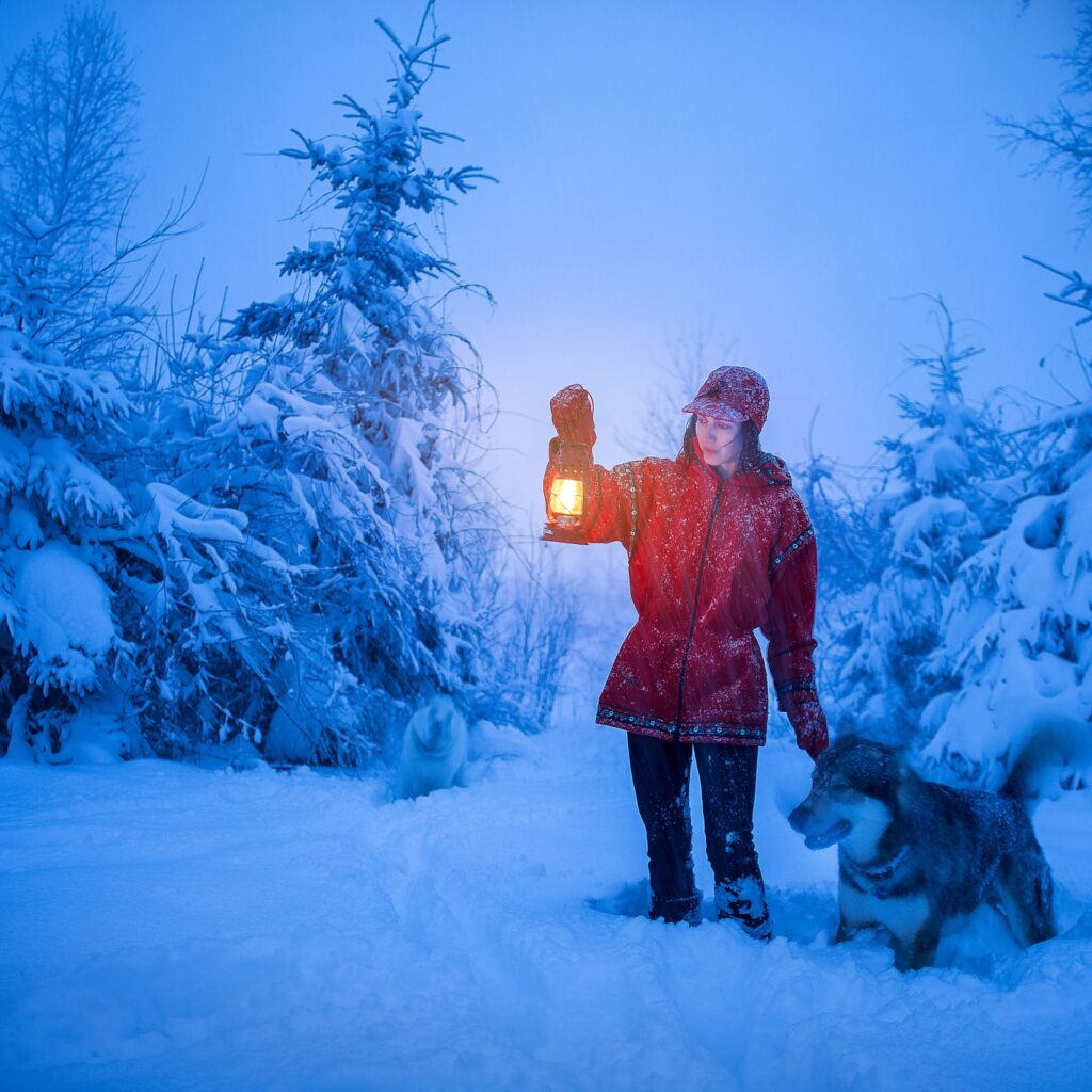 Aurora Acres stands in a snowy wood and holds up a lamp to light the way for her and her wolf.