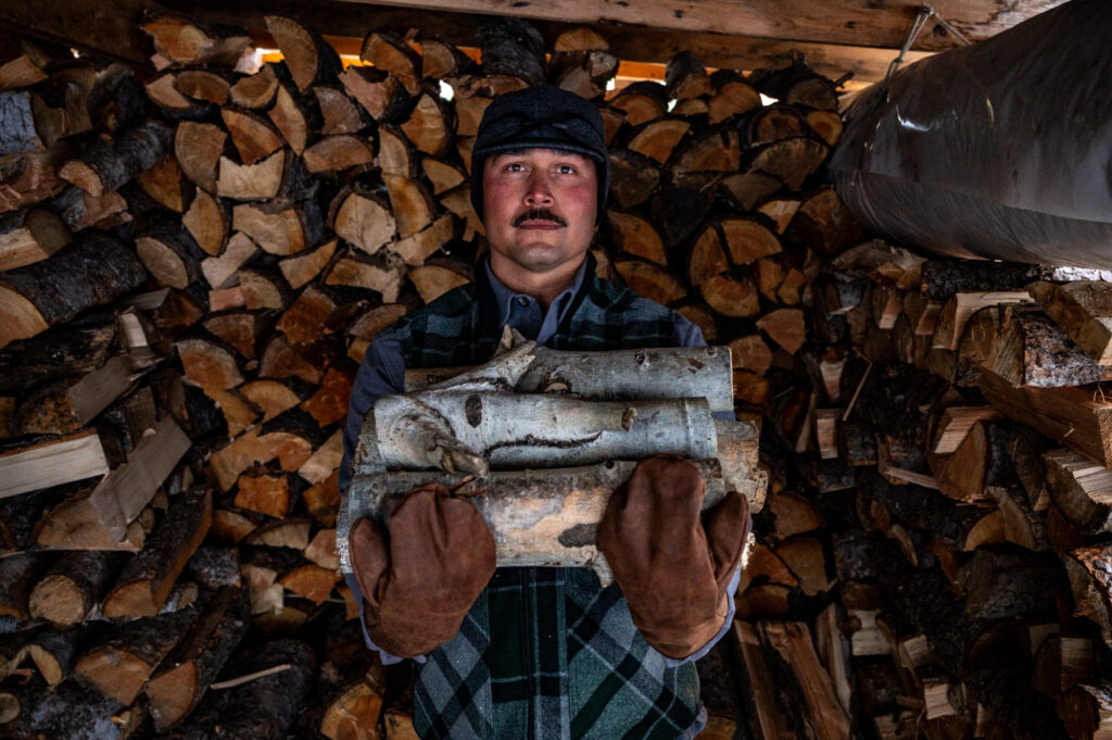 A man holds a bundle of logs in his arms, wearing warm Stormy Kromer outdoor gear and leather gloves.