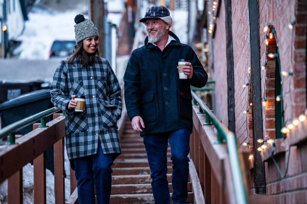 A man and woman walk side by side down an outdoor stairway, holding warm cups of coffee and bundled up in hats and coats.